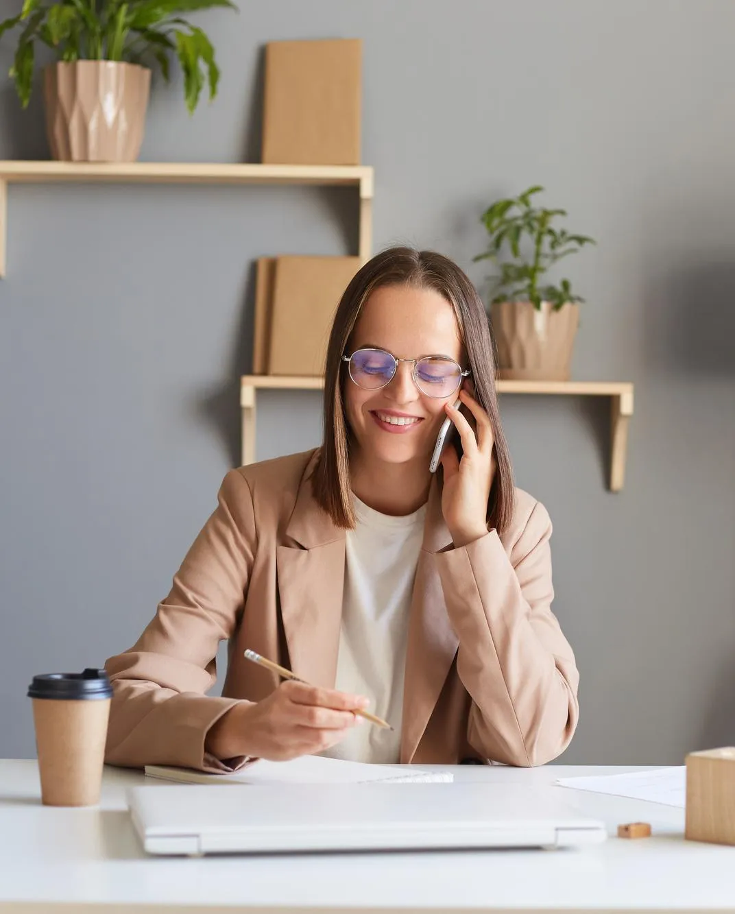 Smiling woman talking on the phone while working at a desk with a laptop and coffee cup.
